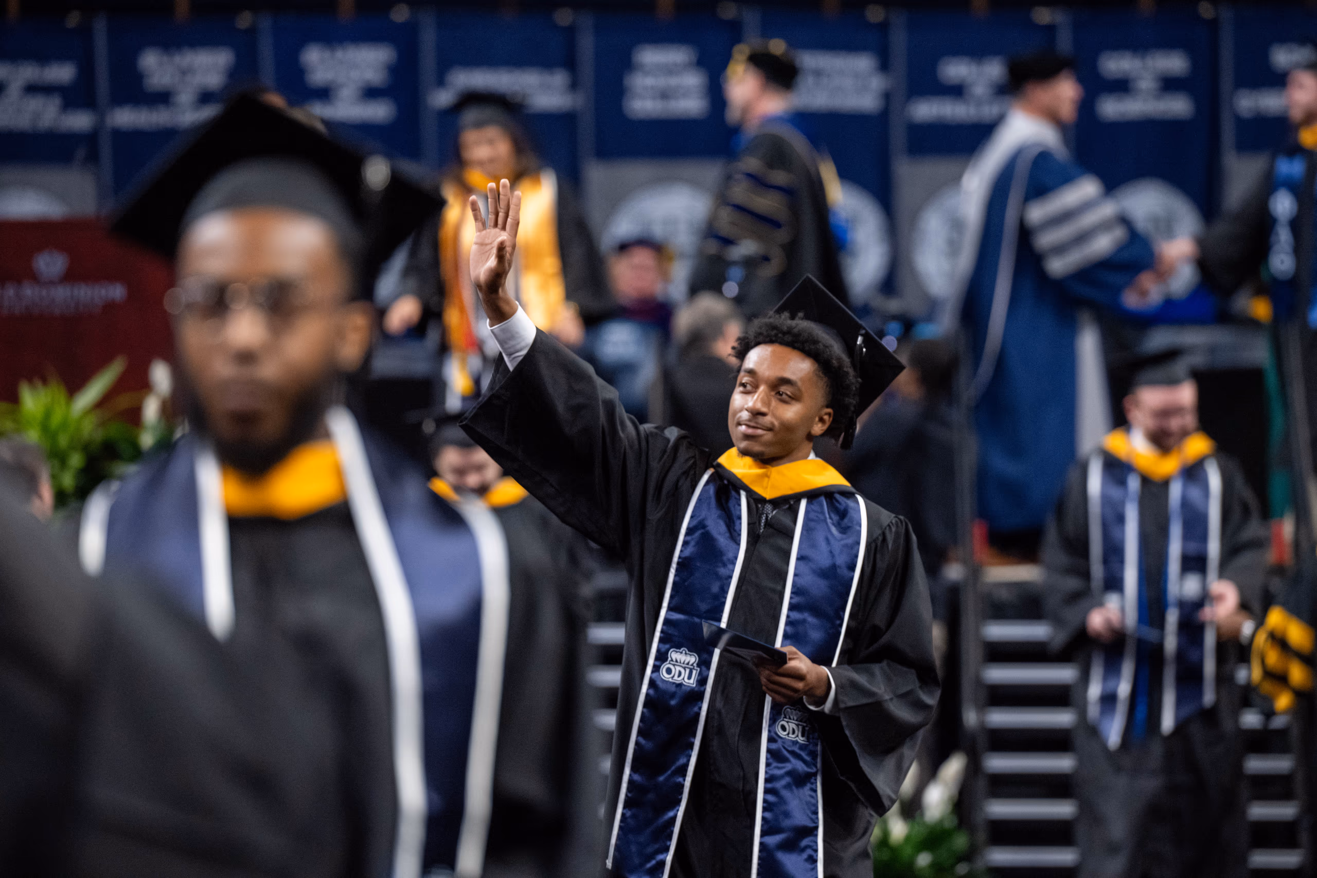 Graduating student at Old Dominion University waving during commencement ceremony, wearing cap and gown with ODU branding, celebrating educational achievements in virginia community.
